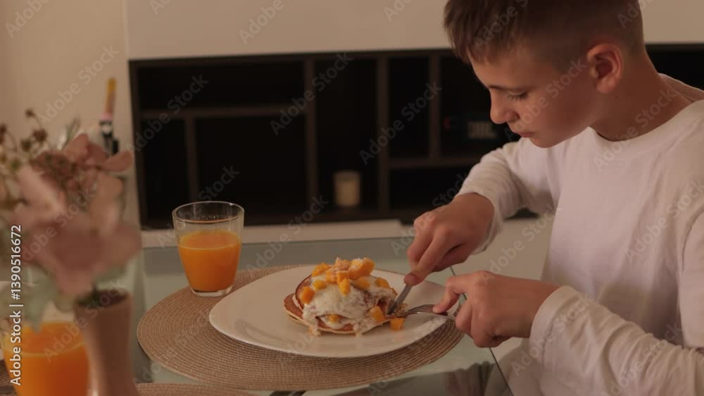 Young boy sitting at glass table, methodically cutting and eating stack of pancakes topped with whipped cream and mango slices, accompanied by glass of orange juice Young boy sitting at glass table, methodically cutting and eating stack of pancakes topped with whipped cream and mango slices, accompanied by glass of orange juice