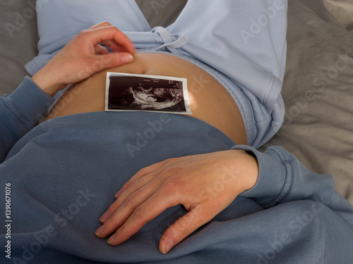 Pregnant woman lying comfortably in blue casual clothes, gently placing an ultrasound scan photo of her baby on her belly, embracing the joy and connection of motherhood