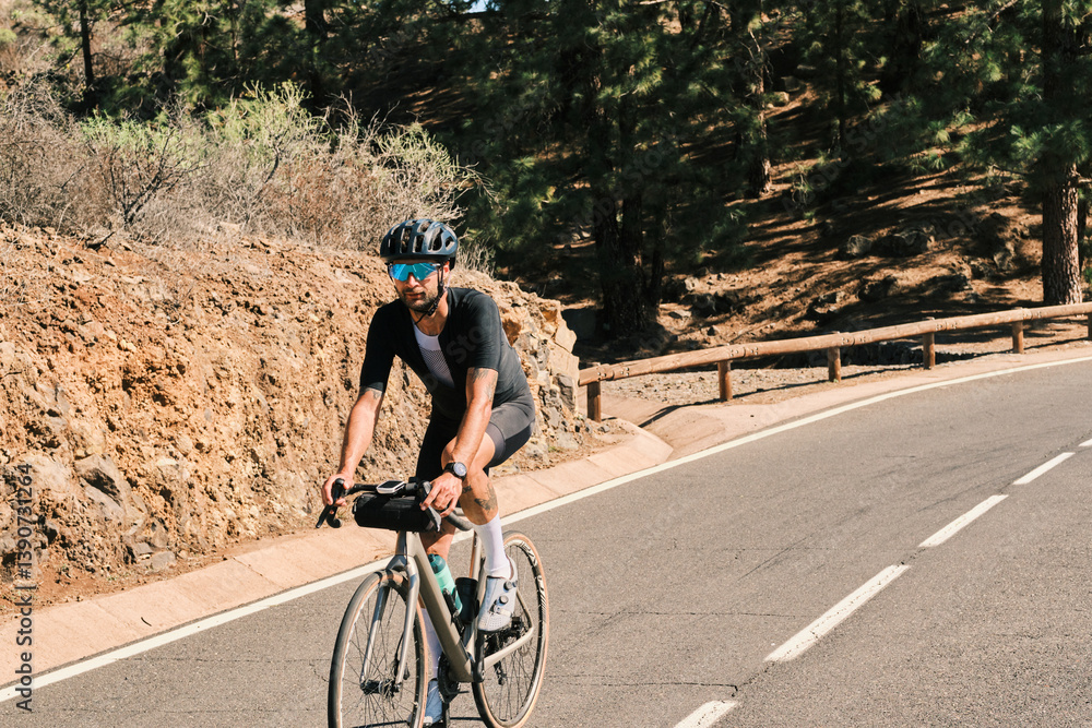 Fototapeta premium A cyclist in black cycling apparel rides a gravel bike along a scenic mountain road. The sun shines brightly, highlighting the stunning natural landscape.