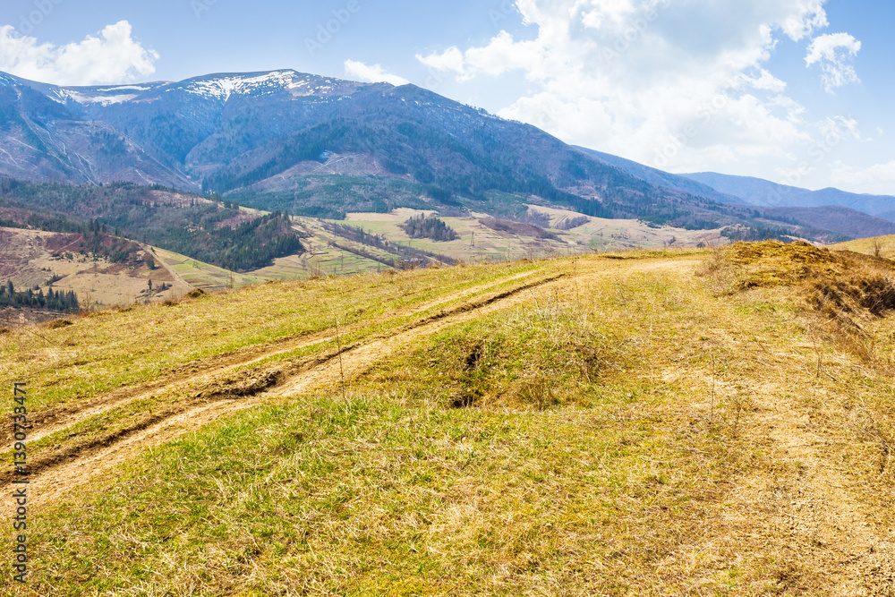 rural road through the hill with grassy meadow. sunny day. carpathian mountain ridge in the distance. alpine countryside of ukraine in early spring. fluffy clouds on the blue sky