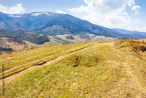 Obraz na płótnie rural road through the hill with grassy meadow. sunny day. carpathian mountain ridge in the distance. alpine countryside of ukraine in early spring. fluffy clouds on the blue sky w salonie