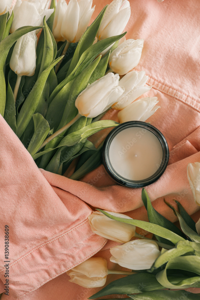 Cozy still life with a burnt candle in an amber glass jar surrounded by white tulips, a pink denim jacket, and croissants in a paper bag. Soft natural lighting enhances the calm, springtime atmosphere
