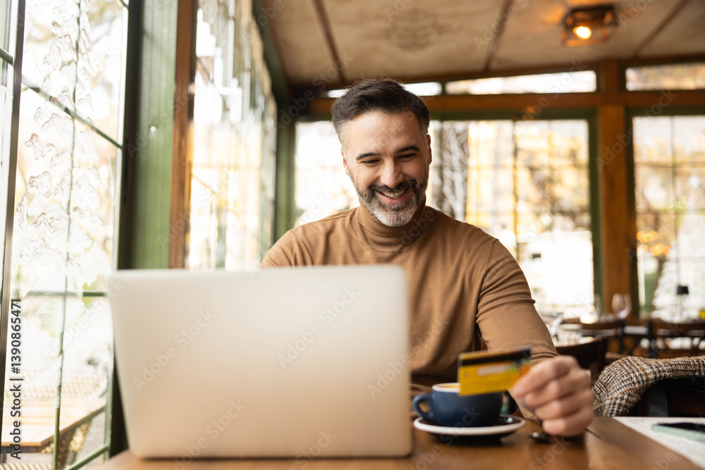 Obraz premium A man smiles while holding a credit card and using his laptop for online shopping in a cozy café setting.