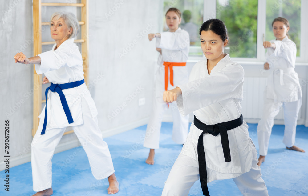 Confident young female martial arts instructor in white kimono with black belt leading karate kata training session for group of women of different ages Confident young female martial arts instructor in white kimono with black belt leading karate kata training session for group of women of different ages