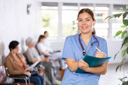 Billede på lærred Polite young female therapist in blue uniform standing in clinic, filling clipboard with medical records