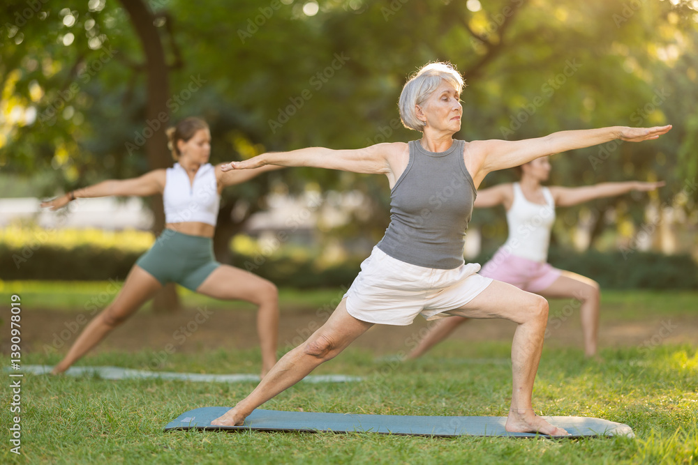 Positive senior woman performing yoga asanas on mat on green glade in serene sunlit summer park, enjoying group session and relaxation in nature. Healthy sports lifestyle for aged adults concept..
