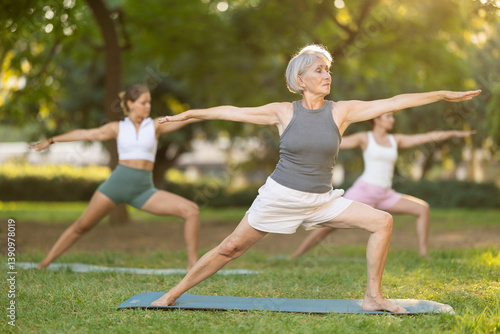 Positive senior woman performing yoga asanas on mat on green glade in serene sunlit summer park, enjoying group session and relaxation in nature. Healthy sports lifestyle for aged adults concept..