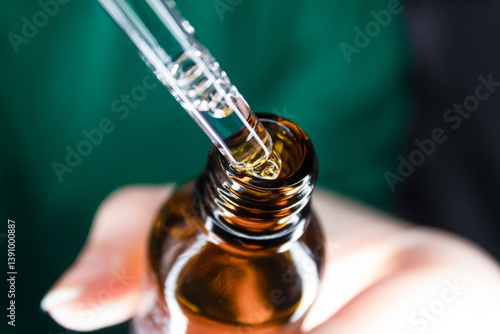 Dropper pipette releasing liquid into a small amber glass bottle. Skincare or cosmetic setting.