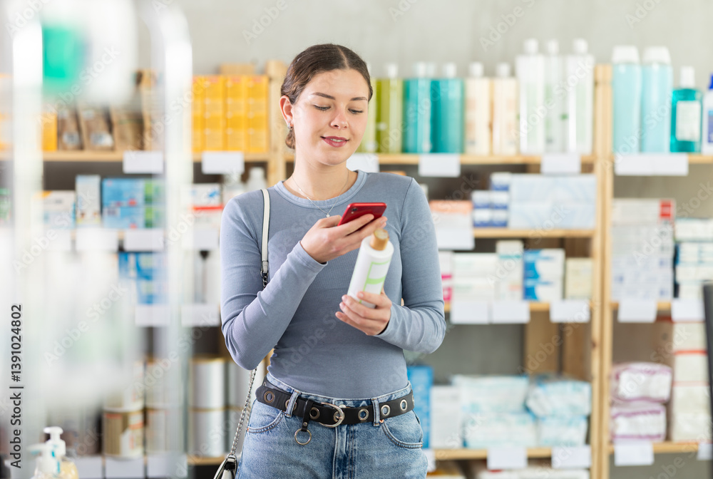 Positive young woman shopping in drugstore for personal care products, using smartphone to scan barcode on bottle of shower gel, checking product information and price. Modern shopping concept