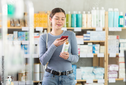 Obraz na płótnie Positive young woman shopping in drugstore for personal care products, using smartphone to scan barcode on bottle of shower gel, checking product information and price. Modern shopping concept w salonie