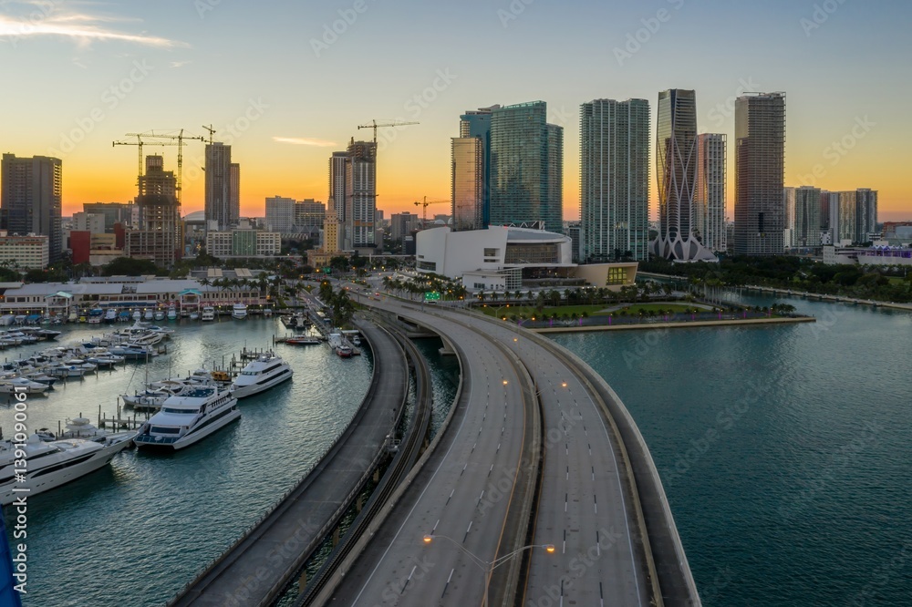 Obraz premium Aerial view of Miami, Florida, USA, at sunset. The photo shows a bridge leading to the city, with a marina full of boats and yachts. Skyscrapers and construction cranes dot the skyline.