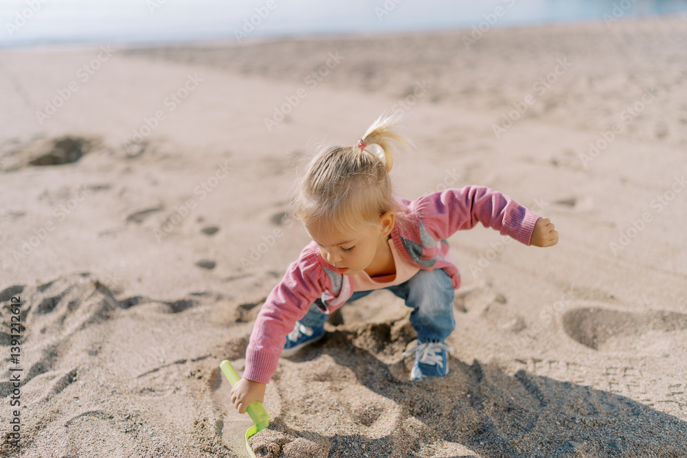 Little girl squats on the beach and digs the sand with a toy shovel with her hand out to the side Little girl squats on the beach and digs the sand with a toy shovel with her hand out to the side