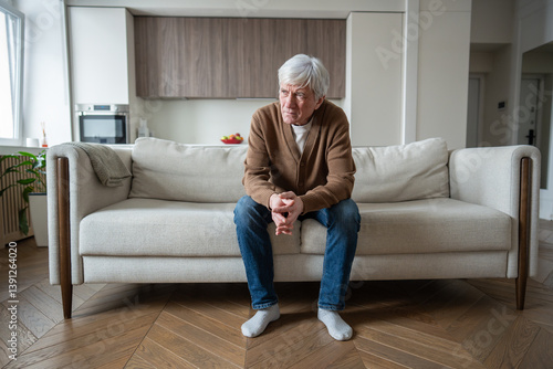 Hunched lonely senior man sitting at home with emotional distress. Old male on sofa looking around, social abandonment, early dementia, chronic illnesses, mental health issues, neglected seniors