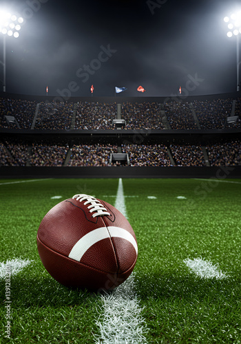 A close-up view of an American football resting on lush green turf, with the stadium lights illuminating the scene and a packed crowd in the background