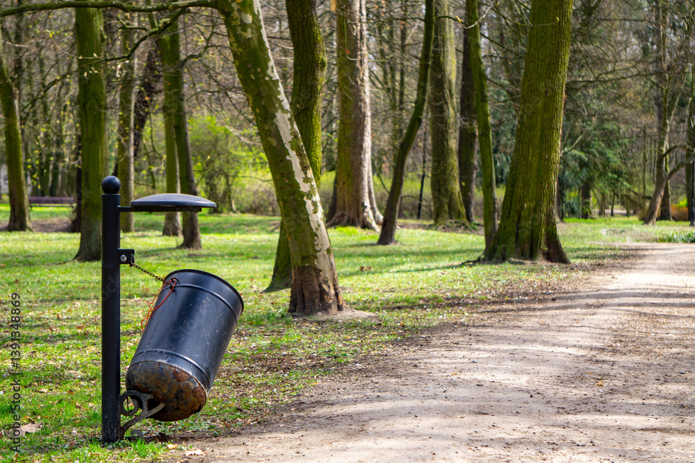 a vandalized, damaged and tilted waste bin mounted on a metal post, located on a gravel path in the park a vandalized, damaged and tilted waste bin mounted on a metal post, located on a gravel path in the park