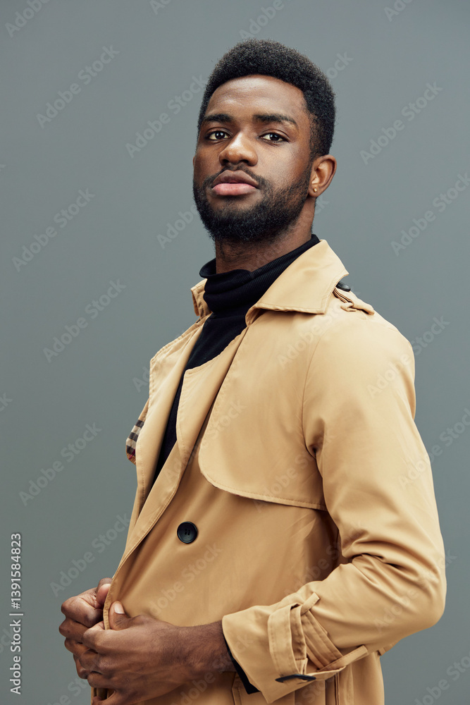 Obraz premium Stylish African man in a beige trench coat poses confidently against a gray background, showcasing modern fashion and self assurance for contemporary lifestyle images