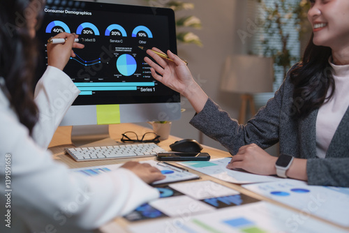 Two businesswomen are collaborating on consumer demographics, analyzing data displayed on a computer screen and using printed reports to gain insights into market trends and customer behavior