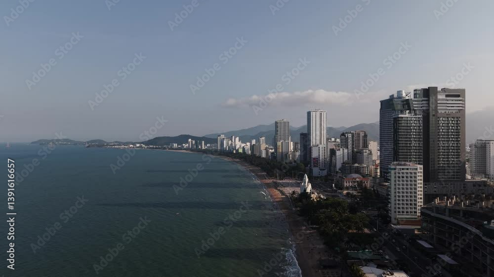 Aerial drone shot flying along a beach in Nha Trang, Vietnam, with towering skyscrapers visible on the right side of the frame. The camera captures the contrast between the sandy shoreline Aerial drone shot flying along a beach in Nha Trang, Vietnam, with towering skyscrapers visible on the right side of the frame. The camera captures the contrast between the sandy shoreline