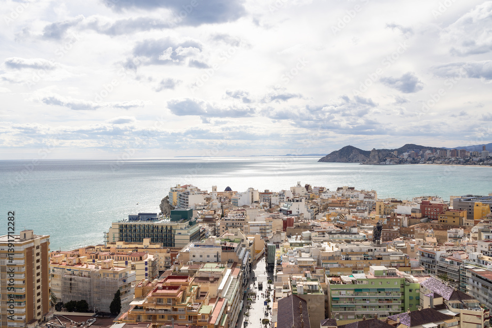 Aerial photo of the Spanish town of Benidorm in Spain showing the old town of the city with the beach known as Platja de Ponent or West Beach Promenade in background as well as mountains and clouds