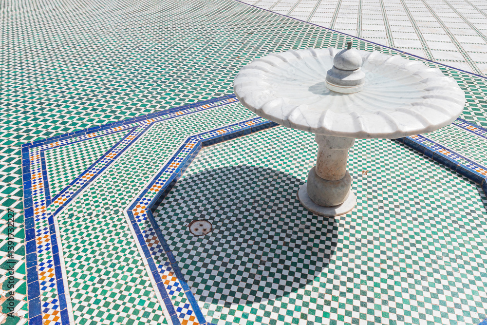 White marble fountain is standing on a checkered mosaic floor with geometric patterns, showcasing traditional moroccan craftsmanship