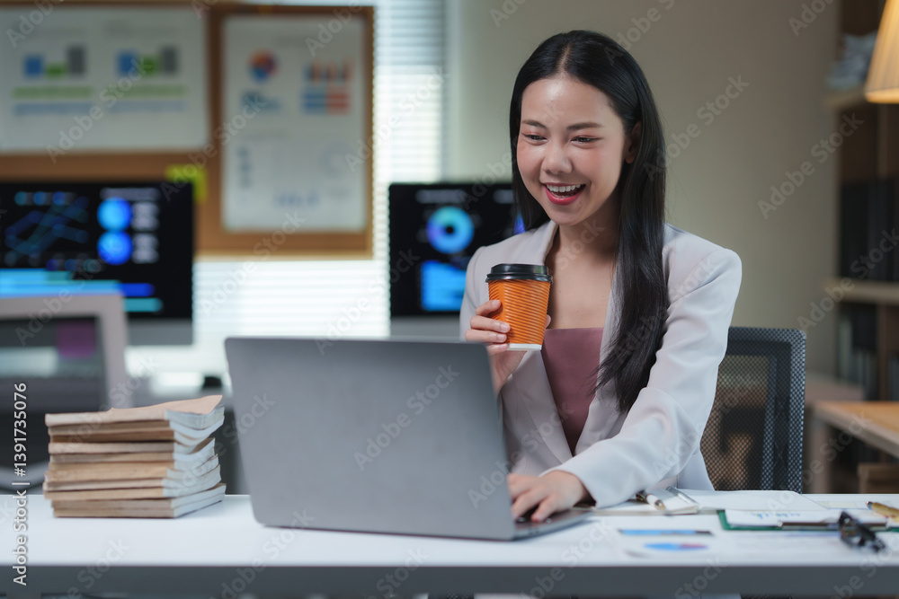 Smiling asian businesswoman enjoying coffee while working on her laptop in the office, radiating happiness from a successful business and effective financial planning Smiling asian businesswoman enjoying coffee while working on her laptop in the office, radiating happiness from a successful business and effective financial planning