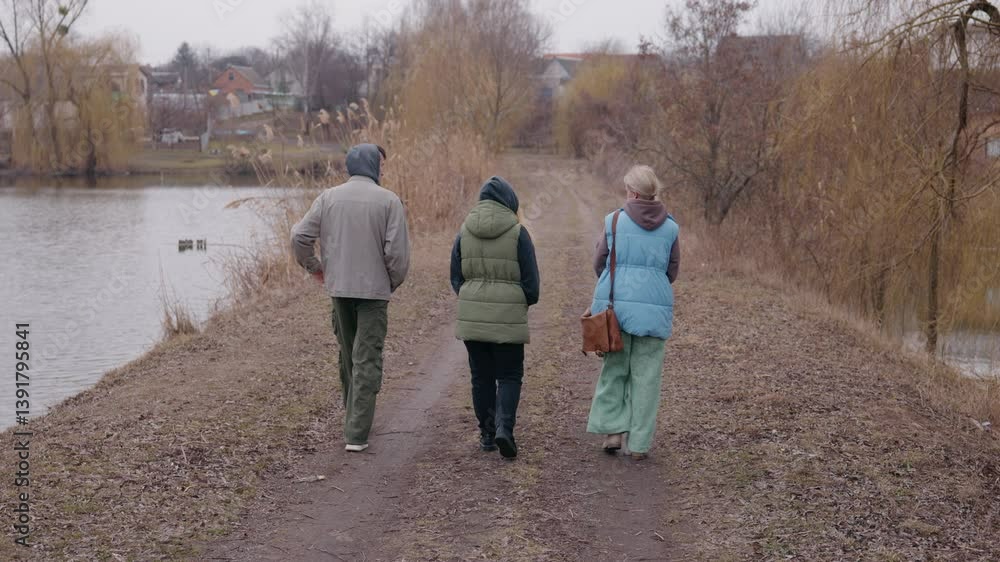 A young couple walks along a dirt path near a lake with their mother on a cloudy spring day. They are dressed warmly, surrounded by bare trees and reeds A young couple walks along a dirt path near a lake with their mother on a cloudy spring day. They are dressed warmly, surrounded by bare trees and reeds
