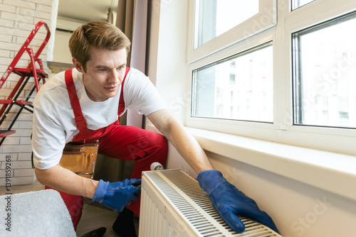 Obraz na płótnie Young handsome professional plumber worker installing heating radiator in an empty room of a newly built apartment or house. Construction, maintenance and repair concept w salonie