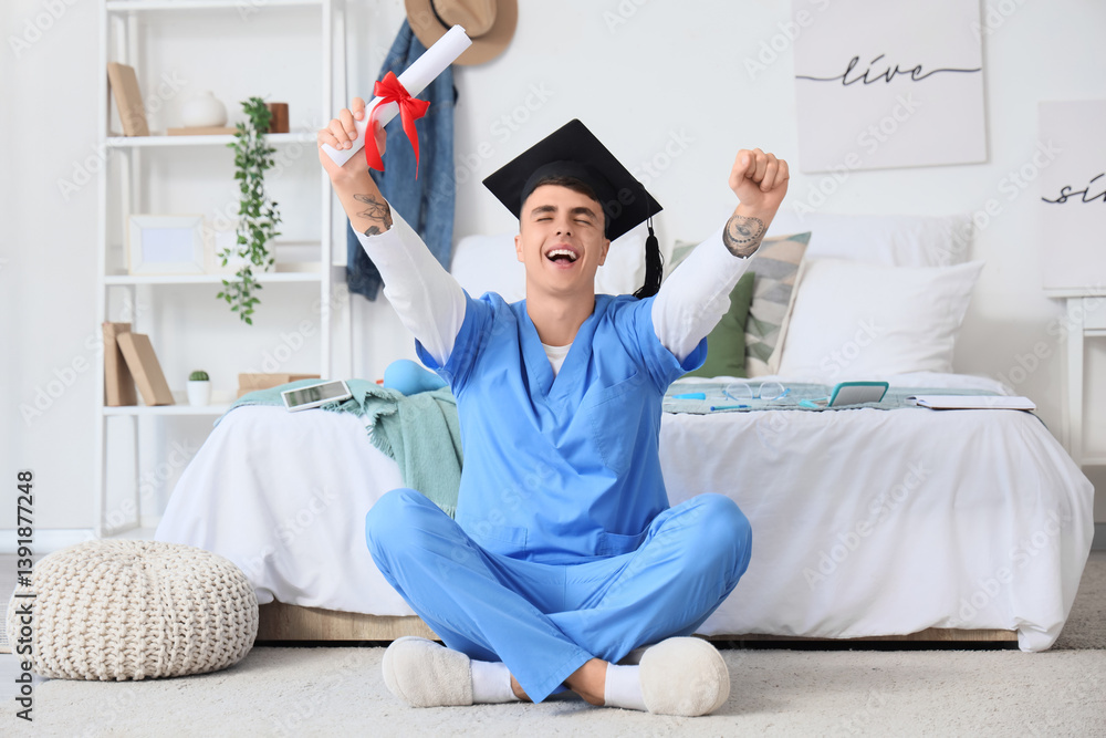 Male medical student in graduation hat holding diploma and sitting on floor at home
