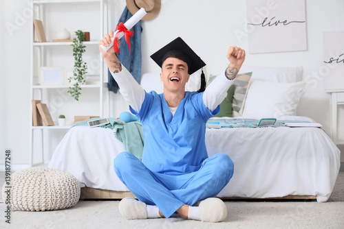 Obraz na płótnie Male medical student in graduation hat holding diploma and sitting on floor at home w salonie
