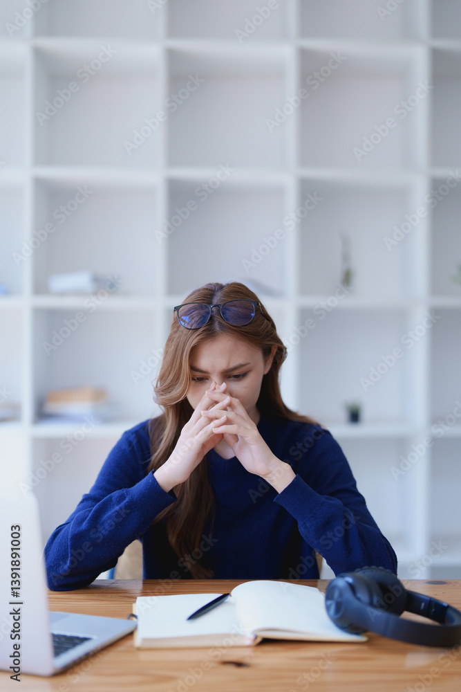 © Jirapong - Stressed young woman overwhelmed by e-learning studies at desk with laptop, notebook, headphones, hands clasped to her face in frustration