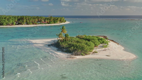 Vivid turquoise coral reef waters surround small, uninhabited island with palm trees, sand shore beach and gentle waves. Serene tropical paradise. Exotic travel getaway background. Aerial static shot