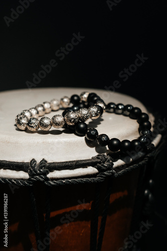 Black and silver beaded bracelets placed atop a traditional djembe drum. The beads' texture and the drum's skin surface create a contrast, merging fashion with cultural music. High quality photo
