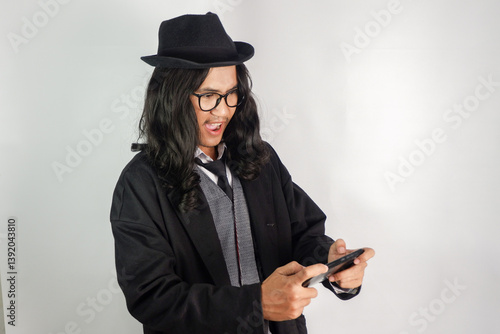 pria asia dengan rambut panjang berpakaian mafia memakai kacamata dan topi bermain game di smartphone dan wajahnya exited