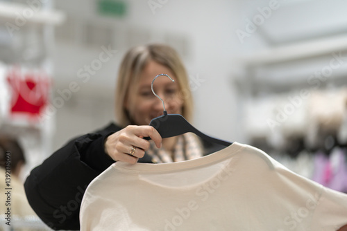 Obraz na płótnie Smiling female customer enjoying a shopping experience in a fashion store, holding a hanger with a stylish white t-shirt while thoughtfully choosing from a variety of apparel w salonie