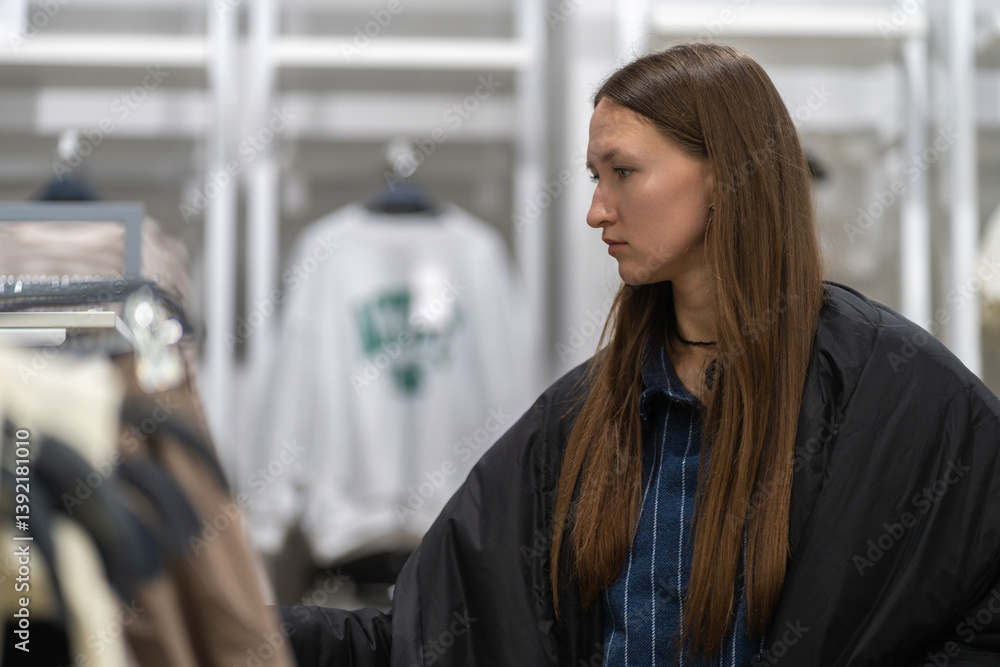 Young stylish woman choosing clothes, shopping in a fashion store, standing near clothes rack, examining garment, making purchasing decision
