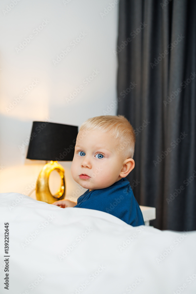 Cute blond toddler boy in blue t-shirt running in spacious hotel room. Toddler boy in comfortable hotel room resting after travel. Child at home sitting on floor