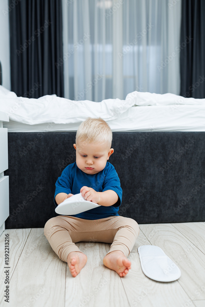Cute blond toddler boy in blue t-shirt running in spacious hotel room. Toddler boy in comfortable hotel room resting after travel. Child at home sitting on floor