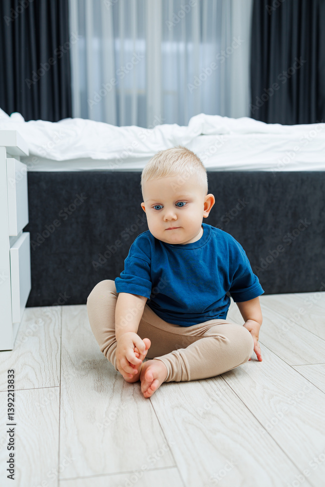 Cute blond toddler boy in blue t-shirt running in spacious hotel room. Toddler boy in comfortable hotel room resting after travel. Child at home sitting on floor