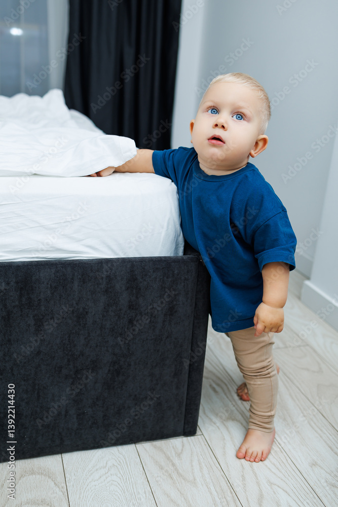 Cute blond toddler boy in blue t-shirt running in spacious hotel room. Toddler boy in comfortable hotel room resting after travel. Child at home sitting on floor
