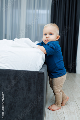 Cute blond toddler boy in blue t-shirt running in spacious hotel room. Toddler boy in comfortable hotel room resting after travel. Child at home sitting on floor