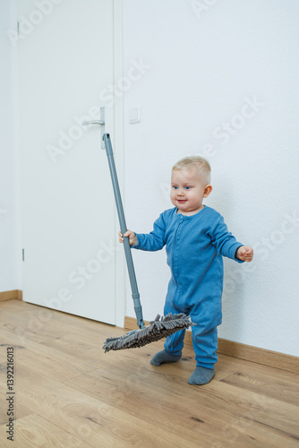 Toddler boy in blue romper cleans the room with a mop. Child washes the floor with a mop. Smiling boy runs around the house and cleans.