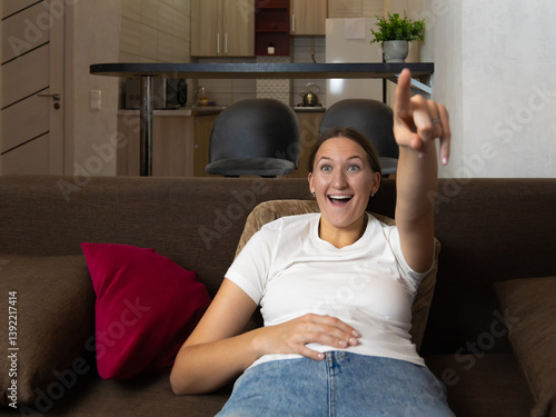 Excited young woman lying on a comfortable sofa is pointing her finger at the television, enjoying a captivating movie or show in her modern apartment