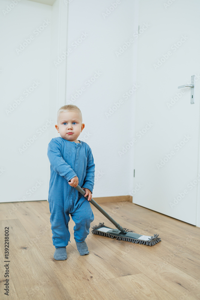 Toddler boy in blue romper cleans the room with a mop. Child washes the floor with a mop. Smiling boy runs around the house and cleans.
