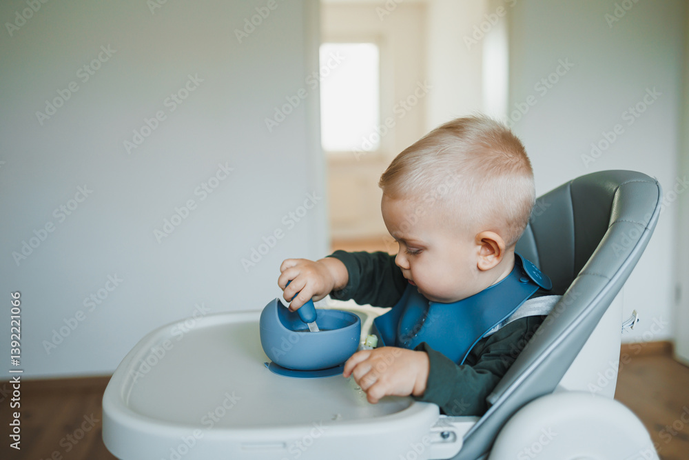 Smiling boy eats soup sitting on a chair. Hungry boy puts food in his mouth. Baby one year and three months old eats independently. Silicone utensils for feeding children. Smiling boy eats soup sitting on a chair. Hungry boy puts food in his mouth. Baby one year and three months old eats independently. Silicone utensils for feeding children.