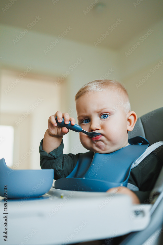 Smiling boy eats soup sitting on a chair. Hungry boy puts food in his mouth. Baby one year and three months old eats independently. Silicone utensils for feeding children.