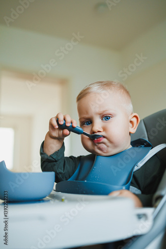 Obraz na płótnie Smiling boy eats soup sitting on a chair. Hungry boy puts food in his mouth. Baby one year and three months old eats independently. Silicone utensils for feeding children. w salonie