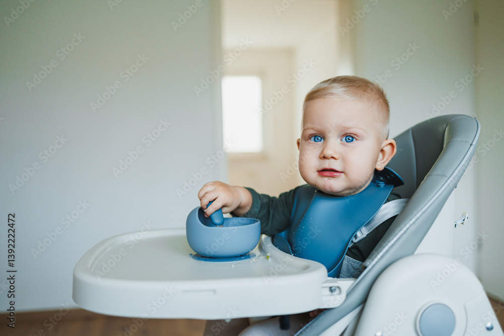A one and a half year old boy sits on a high chair and independently eats soup from a silicone plate with a spoon. Silicone utensils for feeding children. The child learns to eat independently.