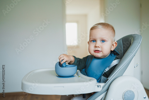 Obraz na płótnie A one and a half year old boy sits on a high chair and independently eats soup from a silicone plate with a spoon. Silicone utensils for feeding children. The child learns to eat independently. w salonie