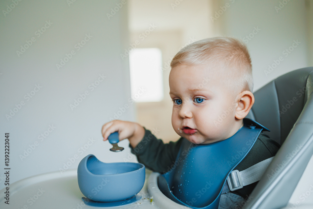 A one and a half year old boy sits on a high chair and independently eats soup from a silicone plate with a spoon. Silicone utensils for feeding children. The child learns to eat independently.