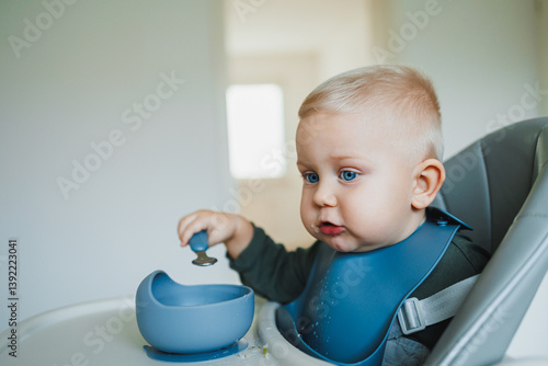Obraz na płótnie A one and a half year old boy sits on a high chair and independently eats soup from a silicone plate with a spoon. Silicone utensils for feeding children. The child learns to eat independently. w salonie